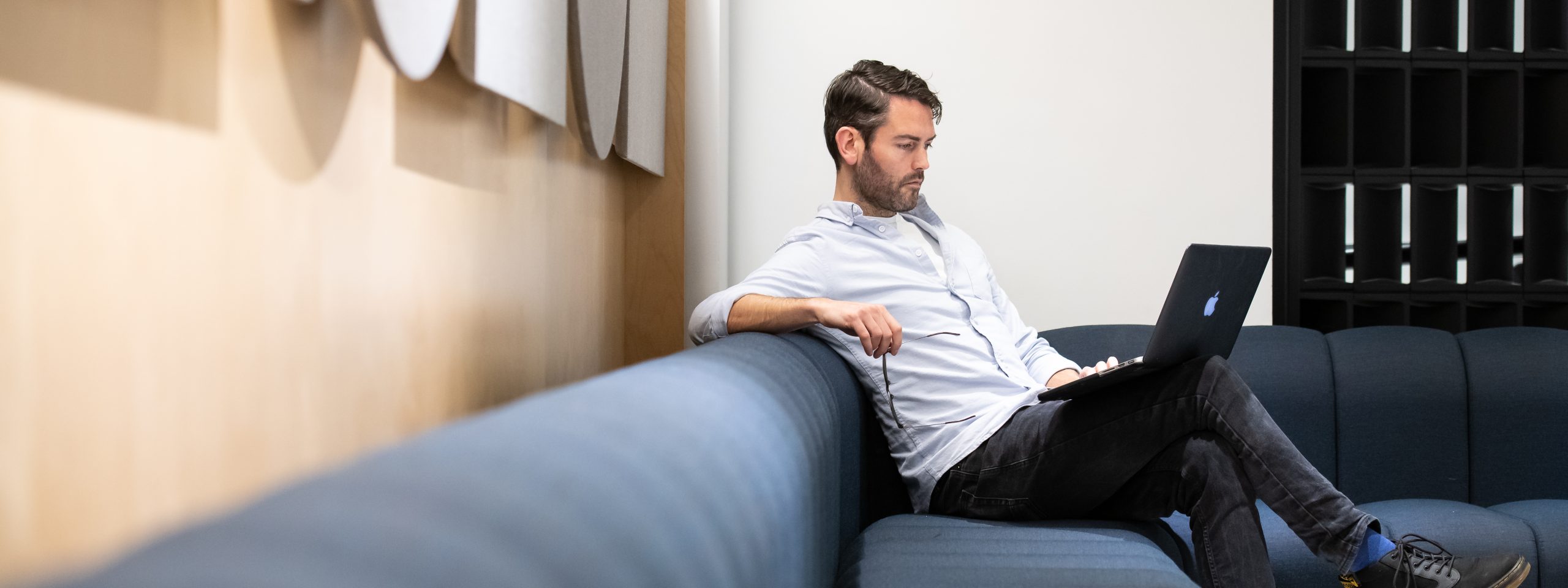 Man in blue shirt reclining on a blue sofa looking at a laptop on his knees.